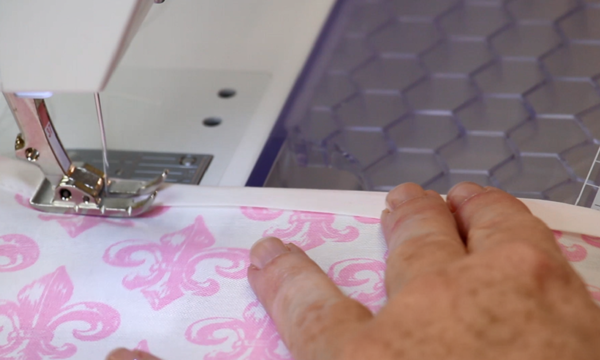 Image shows a close-up of a sewing machine sewing bias tape over the edges of white fabric with pink fleur-de-lis designs.