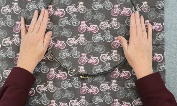 Image shows hands pushing the outer embroidery hoop down on dark fabric with pink and gray bicycle designs that's over the inner hoop.