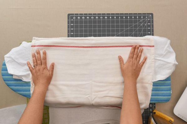 Image shows a press cloth on top of the piece of fusible interfacing atop the cut shirt lying on a small ironing board.