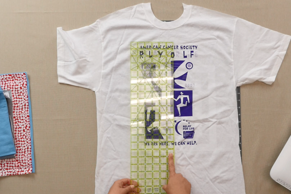 Image shows shirt on a table. Hands are using a ruler to measure the shirt design.