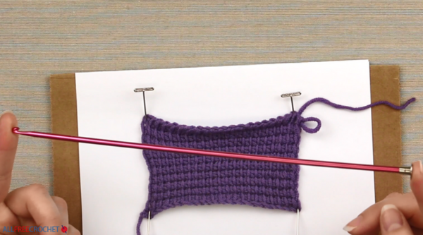Image shows a purple Tunisian crocheted swatch in the background. In the foreground is a Tunisian crochet hook being held on either end my fingers.