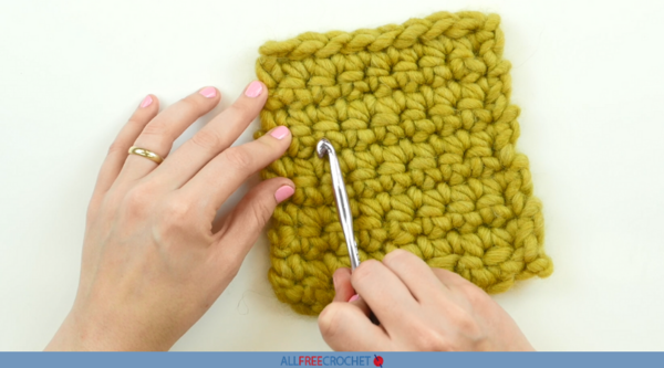 Image shows a swatch of yellow yarn and hands with one holding a crochet hook to show counting stitches.