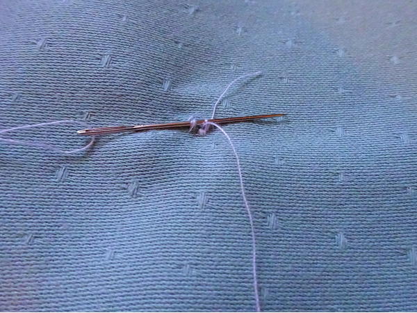 Image shows the back of the sewn button attached to the fabric. The hand sewing needle is being pushed through the loops in order to knot and secure the button.