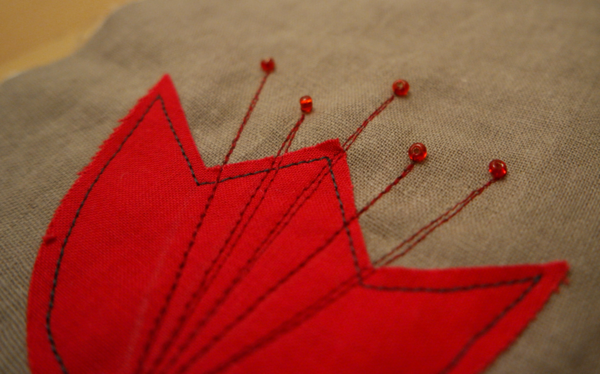 Image shows close-up of a red embroidered flower with four vertical lines added and tiny red beads at the top of the lines, on the cushion. How to Sew the Beads to the Flowers - Step 1