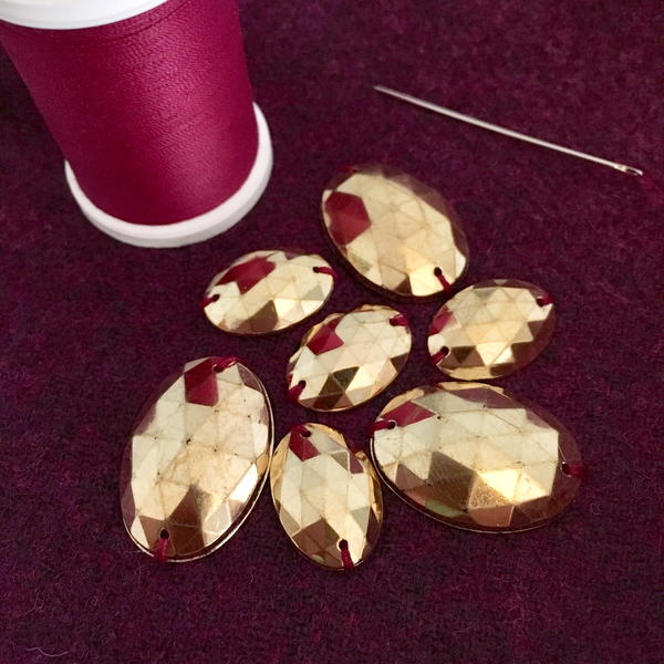 Image shows a spool of thread, large gold sequins, and a sewing needle on a black background.