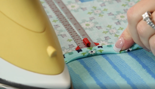 Image shows the edges being ironed up on the fabric top towel piece with a seam allowance ruler to help measure.
