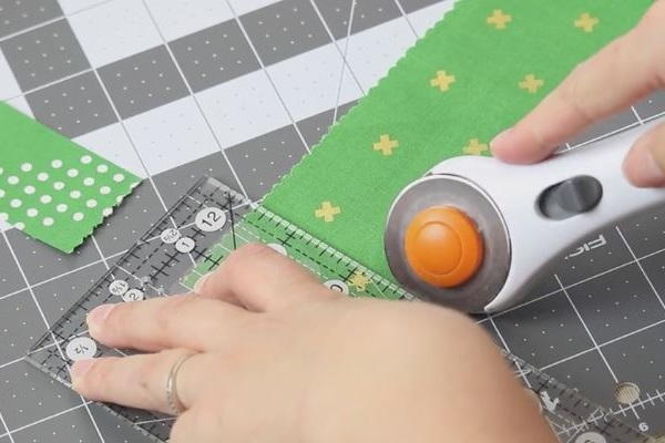 Image shows hands rotary cutting pieces of fabric on a cutting mat using a ruler guide.