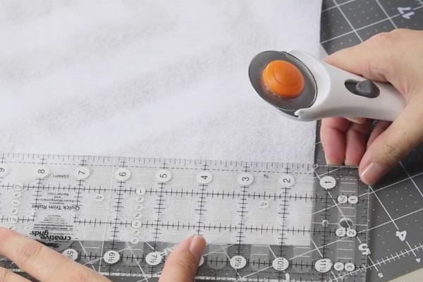 Image shows hands holding a rotary cutter with a piece of fusible fleece on a cutting mat using a ruler guide to measure.