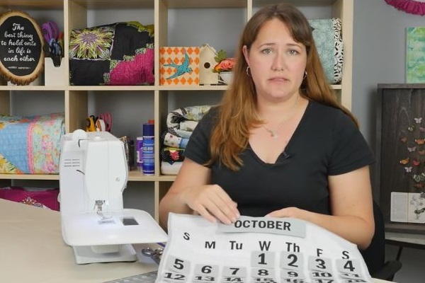 Image shows a woman at a desk with a sewing machine. She is holding the finished quilt calendar.