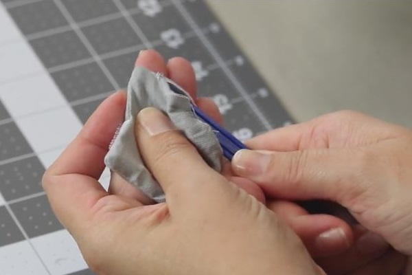 Image shows a cutting board in the background. In the foreground, a hand is holding an object that is pushing the corners out of the sewn fabric square.