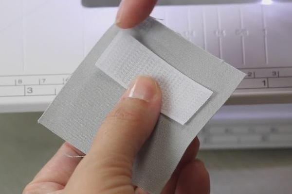 Image shows two hands piecing together a piece of Velcro and a fabric square.