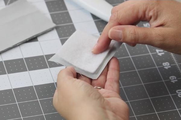 Image shows a cutting board with strips in the background. In the foreground, two hands are piecing together fabric squares.