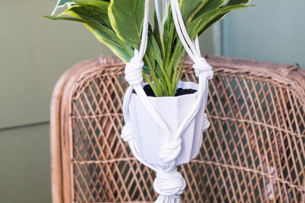 Image shows a wicker chair in the background and in the foreground, there is a white pot with a plant and a white macrame hanger holding it in the air.