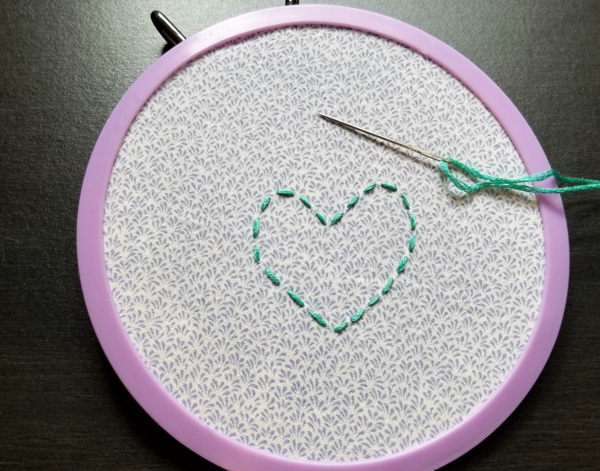 Image shows a purple embroidery hoop with an embroidered heart on the fabric inside. There is a threaded needle resting on the fabric as well.