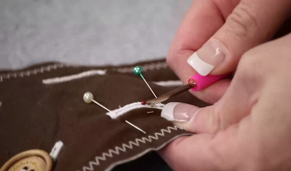 Image shows a brown scrap of fabric with the stitched buttonholes. Hands are using a seam ripper to open the buttonhole with the pins attached.