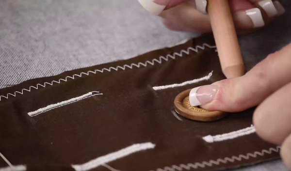 Image shows a brown scrap of fabric with several white stitched buttonholes. Hands are marking a button with a white fabric pencil.