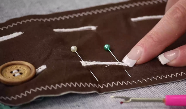 Image shows a brown scrap of fabric with several white stitched buttonholes. Hands are pointing to one buttonhole with pins at each bar tacked end.