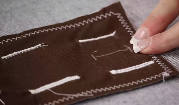 Image shows a brown scrap of fabric with several white stitched buttonholes. Hands are pointing to the white fabric pencil button hole markings.