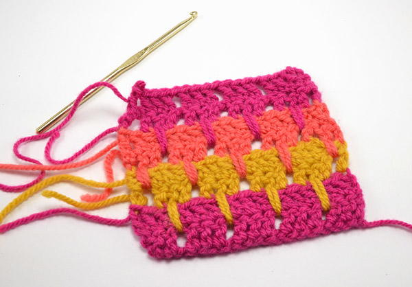 Image shows a crochet larksfoot stitch swatch in pink, orange, and yellow. There is a crochet hook still attached to the yarn strand.