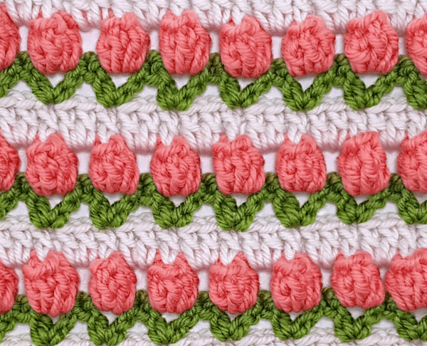 Image shows a close up of a tulip stitch afghan, in white, with pink tulips and green stems.