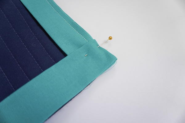 Image shows a close-up of a quilt corner (the quilt is navy, the binding is teal). There is a pin in the fabric at the corner.