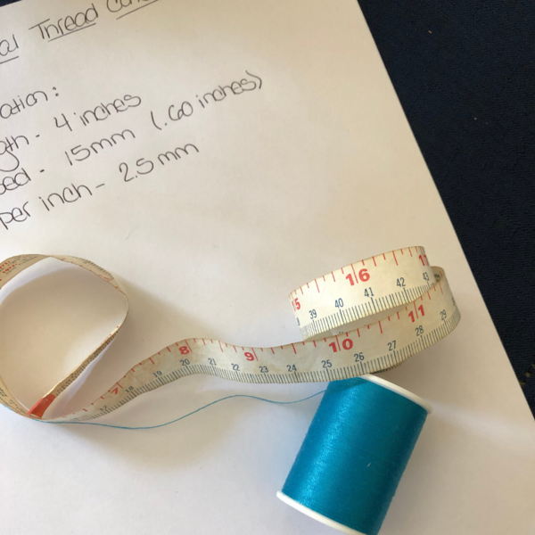 Image shows a piece of paper with thread measurements, a measuring tape and spool of blue thread on top of the paper.