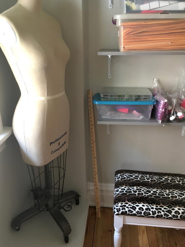 Image shows a corner in a sewing room. There is a body form, an ottoman, and shelves of clear tubs full of sewing materials and fabric.