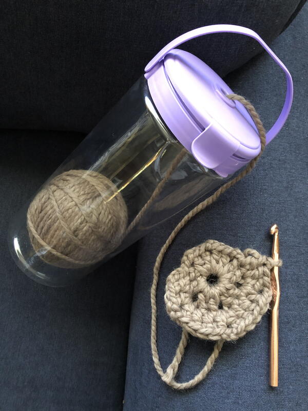 Image shows a plastic water bottle type container with a ball of yarn inside. A strand of yarn is coming out the top hole and on the other end is a crochet circle piece in progress. There is a crochet hook attached to the piece.