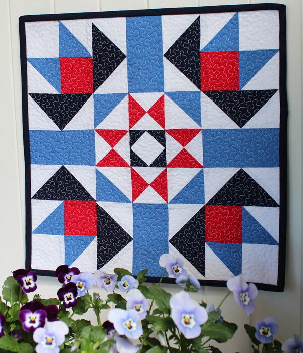 Image shows a barn quilt hanging on a wall with flowers underneath.