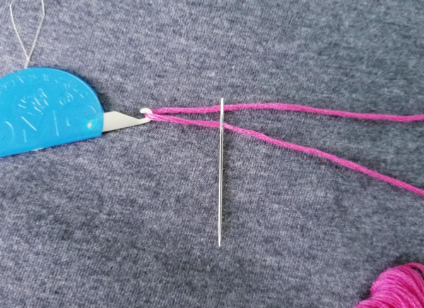 Image shows a needle on a 3-in-1 needle threader with a blue base with pink embroidery floss on the hook on a dark gray background.
