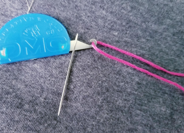 Image shows a needle on a 3-in-1 needle threader with a blue base with pink embroidery floss on the hook on a dark gray background.