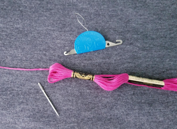 Image shows a pack of pink embroidery floss, a needle, and a 3-in-1 needle threader with a blue base on a dark gray background.