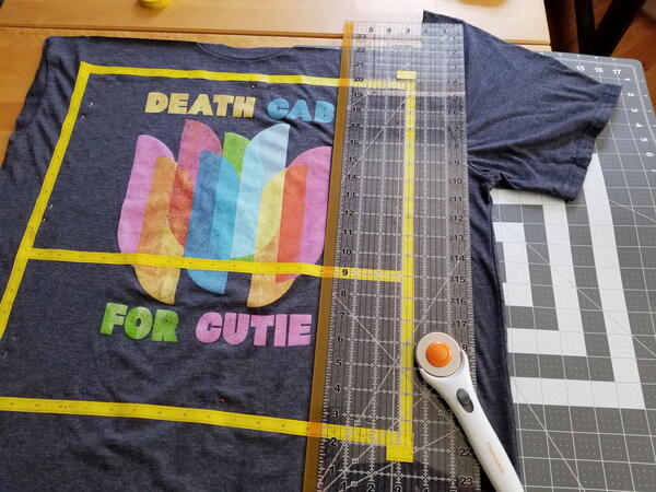 Image shows a shirt with the top removed. It is on a mat with a ruler and rotary cutter laid on top of the shirt.