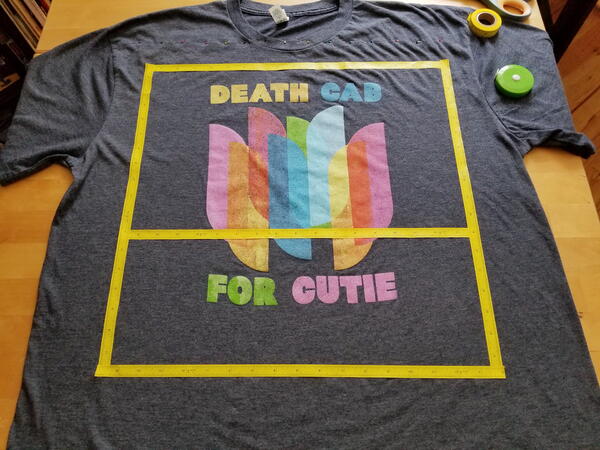 Image shows a t-shirt laid out on a table with yellow ruler tape in a square around the design.