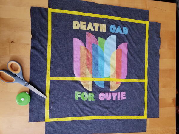 Image shows a cut shirt as a 24" x 24" square on a table with tape marking, and each corner has a square removed. There is a pair of scissors and a tape measure on the table.