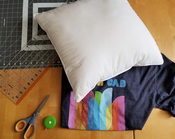 Image shows some of the t-shirt pillow supplies: a self-healing mat, scissors, measuring tape, ruler, pillow form, shirt.