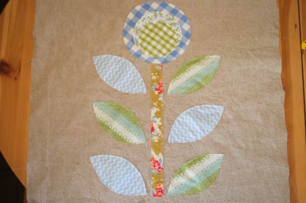 Image shows circle fabric flower, stem, and leaf pieces sewn on the main bag piece, sitting on a wood table.