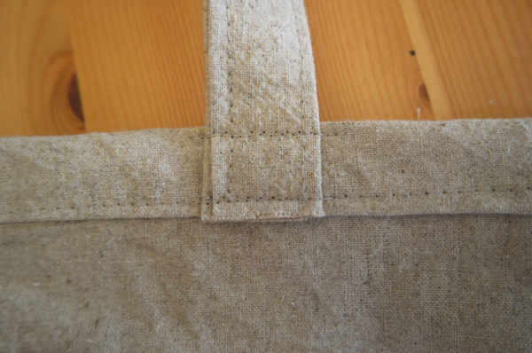 Image shows a close-up of the bag showing the reinforced stitching on the handle base, sitting on a wood table.