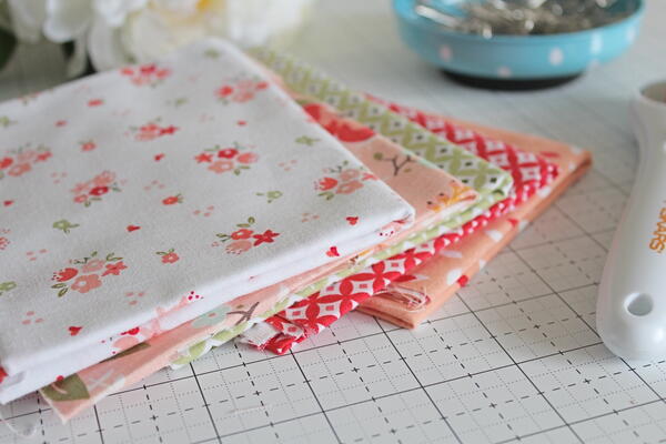 Image shows a stack of fabric on a cutting mat. There's part of a rotary cutter and bowl of safety pins in the image.