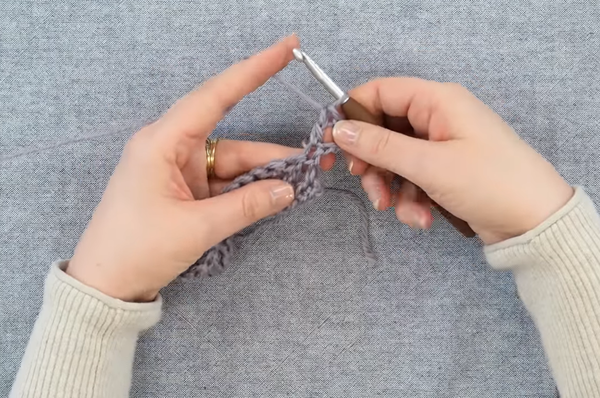 Image shows hands crocheting with a gray fabric background.
