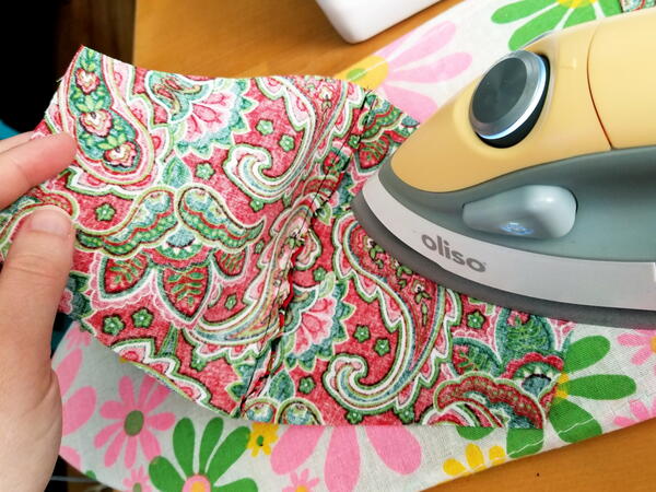 Image shows a hand holding one side of a mask while the other is being ironed on top of an ironing board.