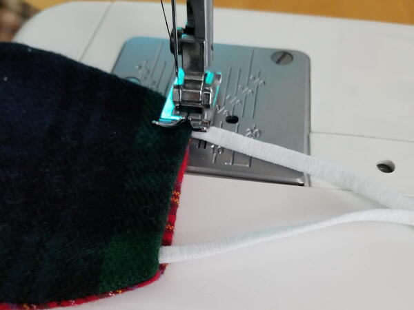 Image shows a close-up of the sewing machine sewing the earloops onto the flannel fabric face mask.