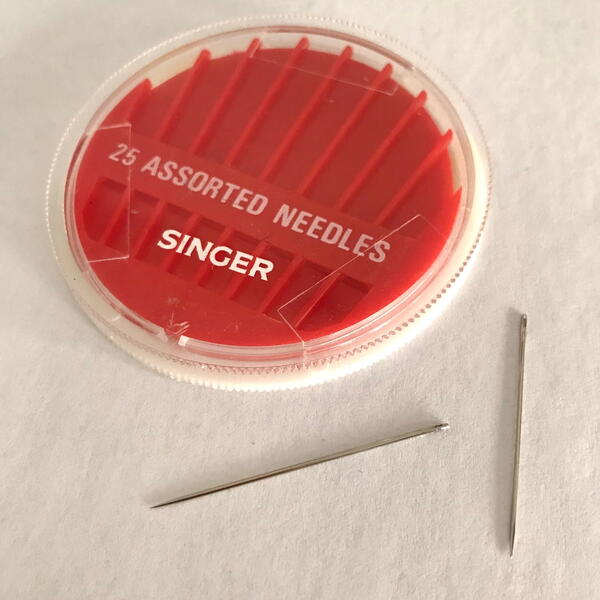 Image shows a package of hand sewing needles with two loose on a white fabric background.
