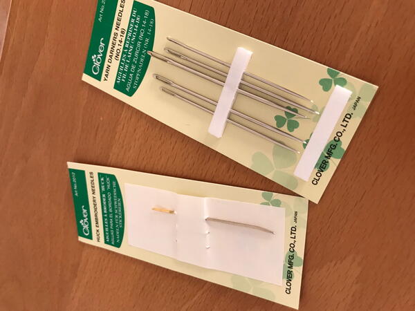 Image shows two packages of various sewing needles on a wood background.