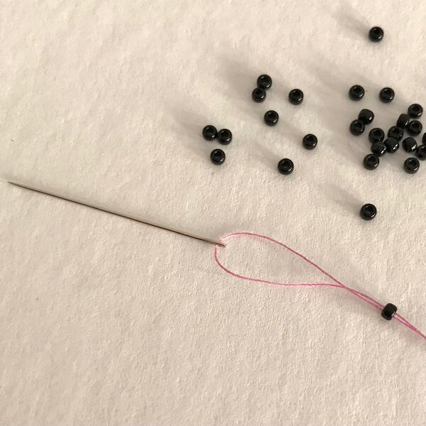 Image shows a threaded hand sewing needle with black round beads on a white fabric background.