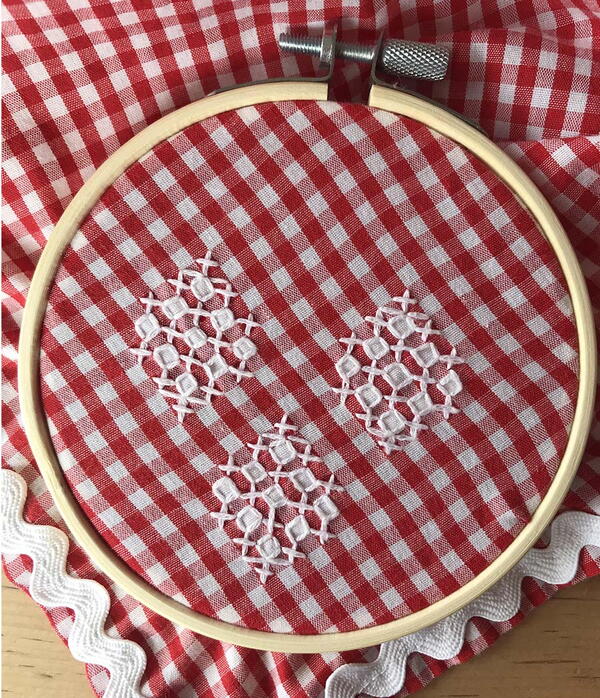 Example of chicken scratch embroidery: red and white plaid fabric with three lacy white stitching sections in a wood embroidery hoop.